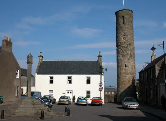 Abernethy Tound Tower, which stands close to the centre of Abernethy, south-east of Perth. The image shows a village square with a tall circular round tower in the far right corner, and a mercat cross in the near left foreground. There are cars parked on the far side of the square and the scene is in sunlight.