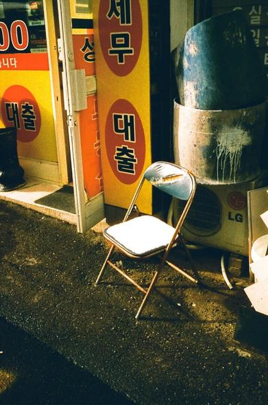 There is a folding chair with a torn top next to the yellow and orange signs, and snow is piled on top of it. The sun is coming down around it.