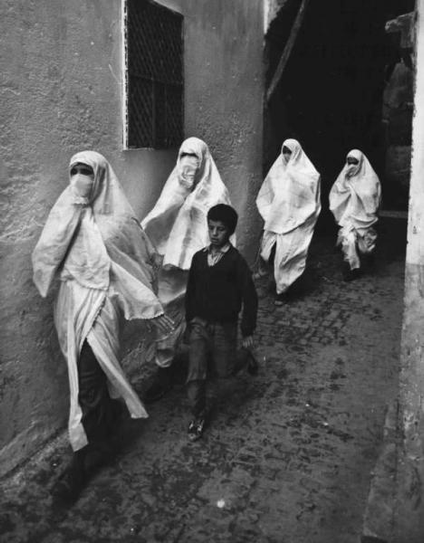 A group of women in white, flowing scarves that cover their heads, shoulders and arms, worn over a white skirt-like part that covers most of the legs (they wear something dark underneath). The lower part of the face is covered with a white niqāb(?). 

Note that the woman on left seems to be wearing a different robe than the 3 following. It has more in common with a bathrobe. Note that this is a film still and that person may be someone in disguise.