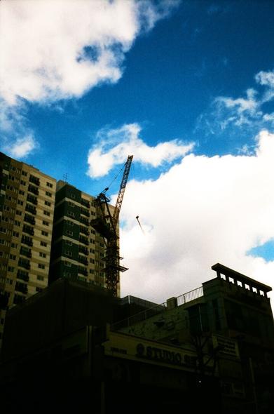 Low building silhouettes and tall buildings, building cranes. Blue skies and white clouds behind them. The pendulum at the end of the crane is swinging like a mobile.