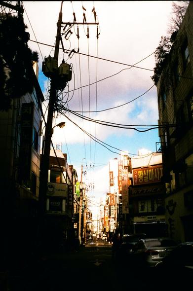 Street view. Nearby buildings and utility poles are shadowed, and the sun is coming down on distant buildings. Above them white clouds and blue sky.