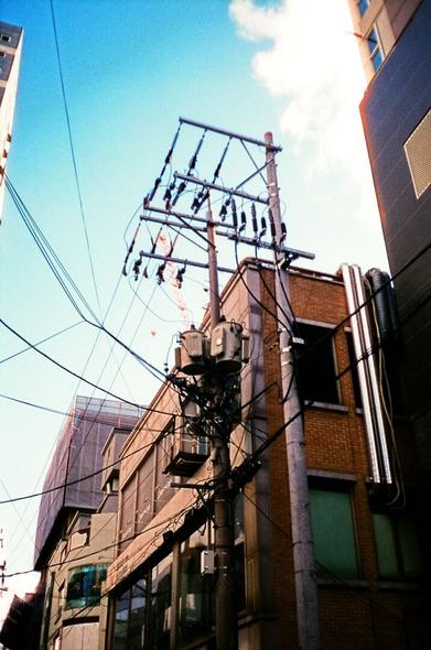 Complex wires stretch in all directions around the utility pole in the middle. Behind it are low buildings and distant construction sites. Behind them are sky blue skies and white clouds.