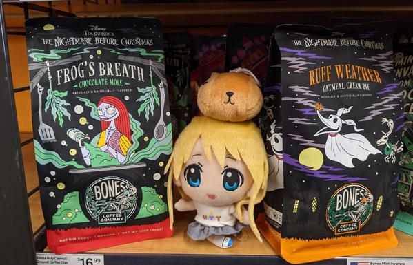 Mugi stands between a bag of Frog's Breath Coffee and Ruff Weather Oatmeal Cream Pie Coffee.