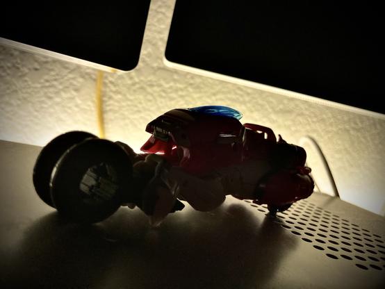 Silhouetted toy motorcycle-like vehicle under a light source with oversized wheels, on a textured surface.