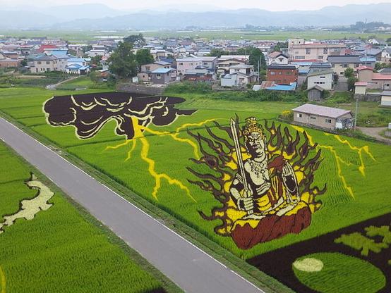 via Wikimedia, Tanbo art, Inakadate, Aomori Prefecture. Acala from the 2012 work “Hibo Kannon and Acala”. Viewed from village administation building: image of Akala, a Buddhist deity, holding a sword and surrounded by flames with a dark cloud and lightning bolt, over several rice paddies in shades of yellow, red, and black