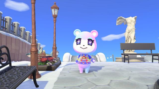 Animal Crossing: New Horizons screenshot. Image showing Judy (pastel blue to purple bear cub with blue / starry eyes) walking along a stone path with some flower beds and street lamps to her left. She is wearing an ugly Christmas sweater with 8-bit Mario in the center of it.