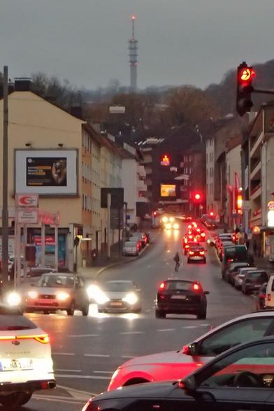 Im Regen: Abendlicher Blick eine ansteigende, städtische Hauptverkehrsstraße hinauf. Autos mit Lichtern fahren über die spiegelnde Fahrbahn. In der Mitte Person, die auf dem Weg über die Fahrbahn den Mittelstreifen passiert. Hinten auf einem Berg ein Funkturm.