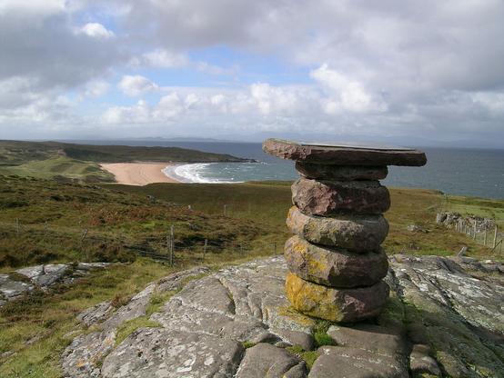 The spectacular and extremely remote shoreline at Redpoint, south of Gairloch in Wester Ross. The image shows a stone viewpoint indicator in the foreground, with a beach in the middle distance facing to the right. There is sea beyond it and then islands on the horizon. The scene is in sunlight.