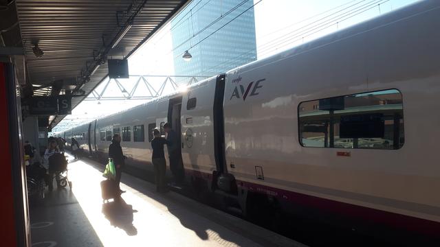 Pociąg Renfe AVE (hiszpański dalekobieżny, wygląda trochę jak nasze pendolino) stoi na stacji w Lyonie. Peron jest "otwarty", widać że pogoda jest słoneczna, w oddali widać wieżowiec.