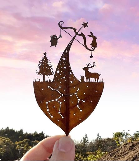 Photo and art. An artfully cut green leaf silhouetted against a blurred sunny background. The artist cuts detailed scenes from foliage leaves. A small frog hangs down on the left side of an archer's bow, while a hare sits on the right side. The bow is strung for shooting and rests on the curved top of a triangular fir tree decorated with small star-shaped holes. A poinsettia is enthroned at the very top. Next to the fir tree is another smaller fir tree on the left and a deer and a mole on the right. The other half of the sheet is the earth/ground beneath the scene. The constellation of the zodiac sign "Sagittarius" is carved into the "earth" in the form of stars. Beautiful filigree work. Incidentally, the zodiac sign Capricorn (the best of all zodiac signs) begins on 22.12.
