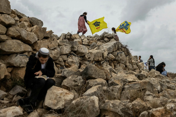 Individuals on a rocky hillside: a person in orthodox Jewish attire reading a book in the foreground, two people holding flags aloft near the top, and others scattered around, some sitting.
