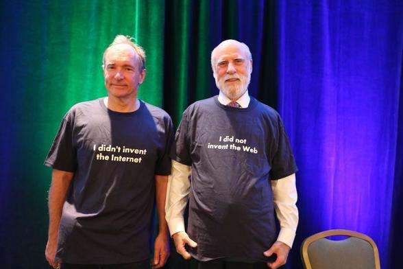 a picture of Tim Berners-Lee and Vint Cerf standing next to each other in front of a green and blue curtain. Tim, on left, is a smiling man with light hair. He's wearing a blue tshirt that says "I didn't invent the internet". Vint Cerf, a smiling man with a white beard, is on the right. He is wearing a white button up shirt and over it, a blue tshirt that says "I did not invent the Web"