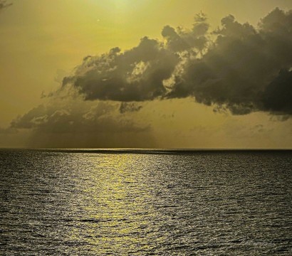 This is a seascape, the Carribean I think though it could be the Gulf of Mexico.  There is dark water as far as the eye can see.  Sky is overcast, but a dim light forces its way through the clouds and reflects off of the ocean. There are low dark clouds floating over the water and in the distance, there is rain.
