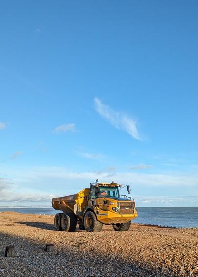 Big yellow work truck carrying shingle from one part of a beach to another. Bright Blue Sky in the background.