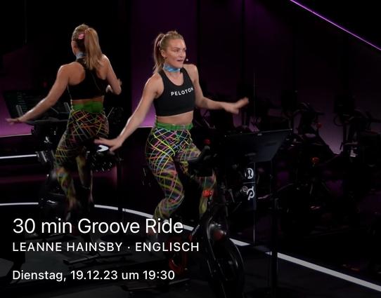 A women exercising on stationary bike in a fitness studio with text overlay announcing a "30 min Groove Ride" class in English, scheduled for a specific date and time.