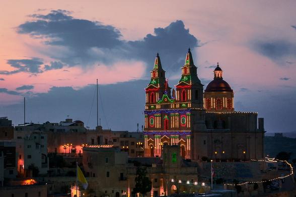 Parish Church of Mellieha in Malta with lights on the building during beautiful pink sky sunset.