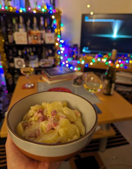 A couch-side view of a bowl containing tartiflette (creamy cheesy potatoes with shallots and pancetta), held up in front of a blurred living-room background decorated with Christmas lights and Christmas decorations.