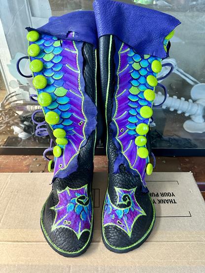 A pair of black leather knee high boots with teal, green, and purple mermaid fins painted on them. A row of green glass buttons runs down the side.