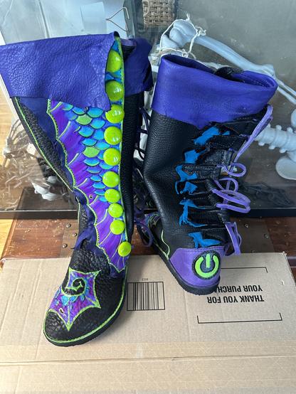 A pair of black leather knee high boots with teal, green, and purple mermaid fins painted on them. A row of green glass buttons runs down the side. One boot is turned around to show the heel has a green power button symbol on it.