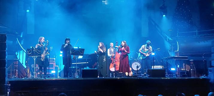 The band "The Unthanks" on stage at Albert Hall, Manchester. Becky and Rachel are centre-stage with hand-helf microphones. The lights cast a blue tinge across the scene.