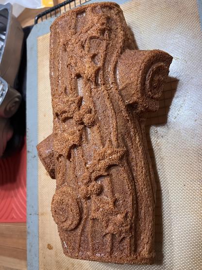 A photo of a brown spice cake shaped like a long log of wood, with two knots from cut “branches” sticking off the sides. The top of the cake has patterns that look like holly berries with spiked leaves attached. The whole cake is resting on a cooling rack with a silicone baking mat between it and the cake. 
Also, you can’t see this, but it smells DELICIOUS and like ginger.