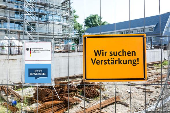 Drei Hinweisschilder an einem Metallzaun vor einer Baustelle. Auf diesen steht: "Wir suchen Verstärkung!" / "Bundesministerium für Wohnen, Stadtentwicklung und Bauwesen" / Jetzt bewerben!".