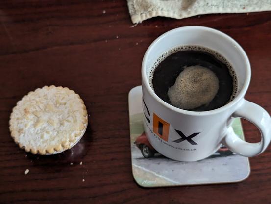 Photo of a mince pie and a mug of black coffee on a table viewed from a high angle. Mug has CIX written on it.