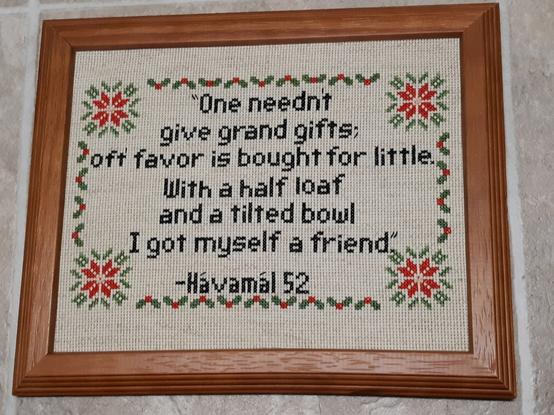 An eight by ten inch cross stitch piece in an oak frame. It has text from verse fifty two of the Havamal bordered with poinsettias and holly. It reads: One needn't give grand gifts; oft' favor is bought for little. With a half loaf and a tilted bowl, I got myself a friend.