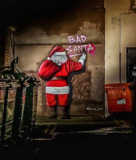 Streetartwall. A Santa Claus figure was sprayed/painted on a street wall in a dark backyard with garbage cans. Santa Claus can be seen from behind, carrying a red sack full of spray cans over his shoulder. He is spraying the words "Bad Santa" on the wall of the house with a spray can.