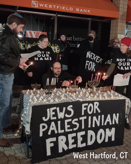 A table lit with menorah and tea candles reads JEWS FOR PALESTINIAN FREEDOM in white text on black, and is surrounded by men and women, some in kippah, wearing black shirts with white text which reads NOT IN OUR NAME holding a banner that says NEVER AGAIN FOR ANYONE. Another banner reads CEASEFIRE in flames with FREE PALESTINE beneath a menorah. The watermark in the corner says West Hartford, CT.