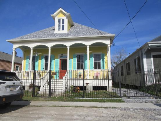 5 bay wooden house, raised on piers a meter above ground, with a central front stairway leading via covered front porch to the central front door, flanked by pairs of windows, all with aqua blue shutters against yellow wall. A single dormer window is above the central door.