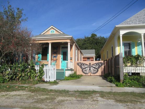 Single story wooden house, 2 bay "shotgun" style with wider projection towards the back right. There is a driveway on the right side with a wooden gate, painted with a large butterfly. Smaller butterfly designs adorn the front of the house.