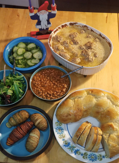 Photo of Swedish meatballs, Boston beans, a huge loaf of bulle, cucumber salad, green salad, & hasselback potatoes all being watched over by a tomte candle holder.