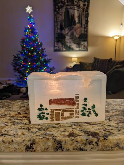 A blocky rectangular candle sits on a kitchen counter. It has a cozy winter scene carved and painted on it. It's glowing and behind it you can see a lit up Christmas tree in a living room.