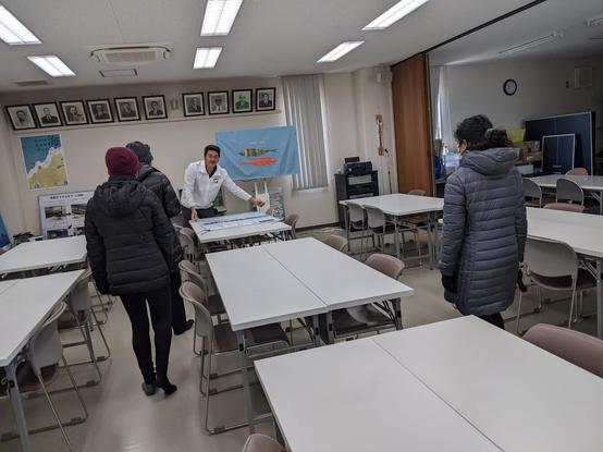 A room with many long tables and chairs - a person is placing a poster on a table as 2 kids and a parent are going up to the table with the poster