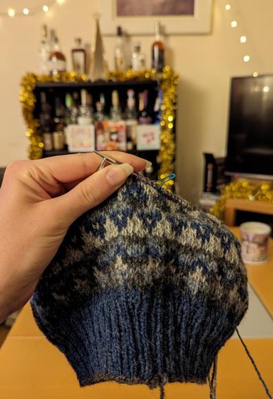 A blue and grey colourwork hat knitting WIP being held up against a living-room background. The hat is on circular needles and about 2/3 knitted.