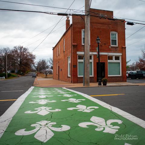 Crosswalk painted green with white fleur-de-lis in front of Old, two-story brick building.