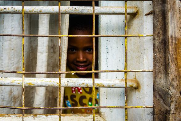 A near view of a white barred shanty window with a young boy's hazy grinning face peering from a dark interior.