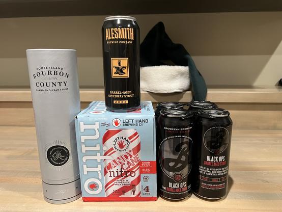 A collection of fine beers - left to right, a tall silver cylinder hold a $40 bottle of Goose Island Bourbon County Rare Eagle barrel-aged stout, a 4 pack of LeftHand Candy Cane Nitro stout (complete with red and white candy cane stripes on the cans), a dark black can of AleSmith Barrel-aged Speedway stout, and a 4 pack of BlackOps barrel-aged stout also in black cans