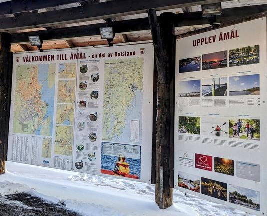 Photo of an informational map poster at the side of the road, with the title "Välkommen till Åmål", meaning "Welcome to Åmål". On the right is another information poster with photos of various things to experience in Åmål with the title "Upplev Åmål", meaning "Experience Åmål".