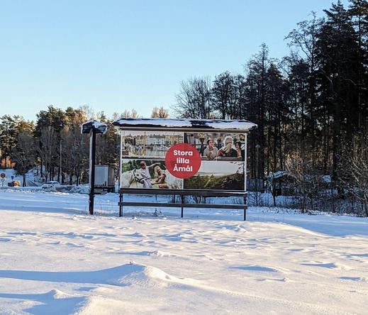 Photo of a big sigh standing in a snowy area with trees in the background. On the sign there's four photos of summer activities in the area, with a big read circle label with the text "Stora lilla Åmål", meaning "big small Åmål".