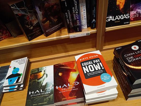 Eine Bücherauslage in einer Buchhandlung. Im Hintergrund stehen einige Science Fiction Romane. Im Vordergrund liegen die Romane "Per Anhalter durch die Galaxis", "Halo - Stiller Sturm", "Halo - Oblivion", das Sachbuch "Equal Pay Now!", der Roman "Die drei Sonnen"