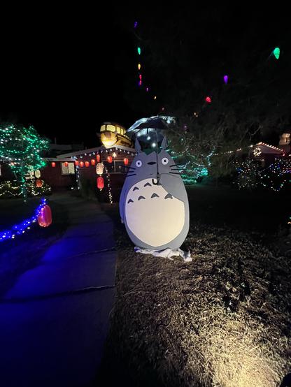 Totoro in the foreground with colorful outdoor holiday lights and decorations in the background during nighttime. Catbus on roof