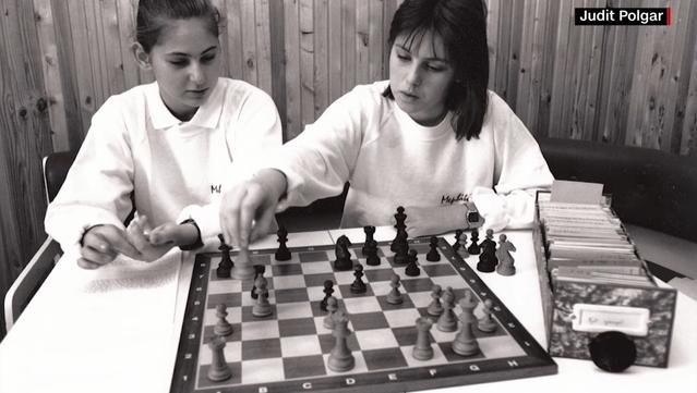 Black and white photo of two female teens sitting on the far side of a chessboard. Sitting next to the board is a tray about as long as the board which is full index cards.