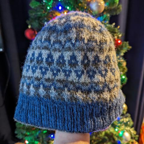 A blue and grey stranded colourwork hat, with geometric shapes made of MC dark blue and CCs dark grey, light grey and light blue. It is being held up in front of a Christmas tree.