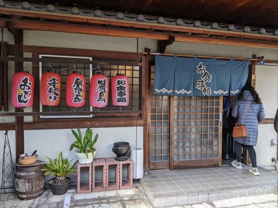 Outside of a restaurant in the daytime - 5 red lanterns are outside the window to the left of the sliding door which has short blue curtain in front with the name of the restaurant in Japanese (Kadoya)