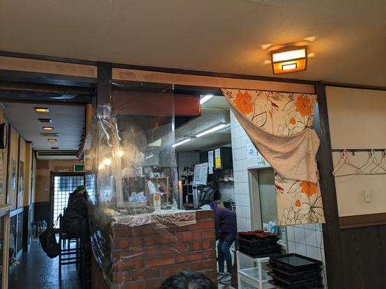 Inside of a small restaurant. The kitchen is behind a counter that leaves a narrow passage between the seats and wall behind. The picture is taken looking at the sliding door  entrance from a table space past the counter