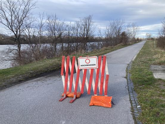 ein Scherengitter steht halb über einer schmalen Straße oder einen Radweg an Flussufer. Hoher Wasserstand in Fluss links vom Radweg. Links und rechts Gras und winterliche Bäume, die links stehen im Wasser.
