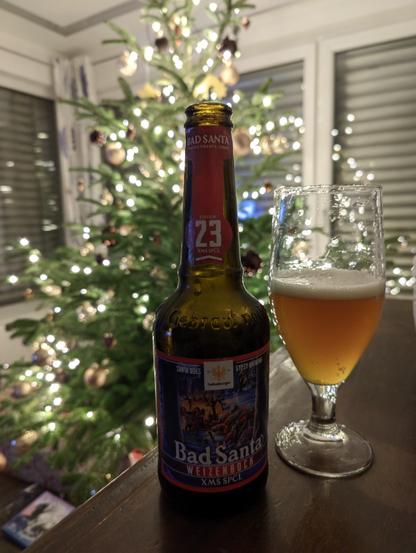 Bad Santa ein Weizenbock steht auf einem Kästchen, daneben ein damit halb gefülltes Bierglas, im Hintergrund ist ein Christbaum