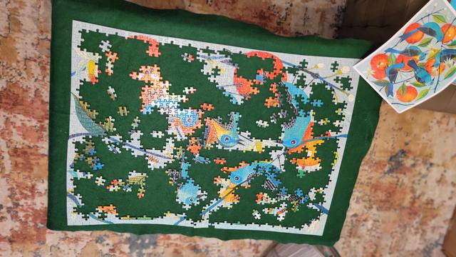 Photo from above of a partially finished 1000-piece puzzle with the shapes of birds, big oranges lightly speckled with white spots, white flowers striped with pale turquoise, buds, leaves on two shades of green, and bees on a gray background.